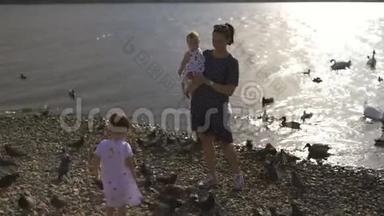 年轻的母亲带着她的小女儿在河边喂天鹅和小鸭子，小鸟在河边吃面包，穿着点缀的裙子